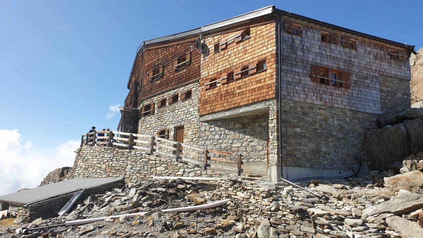Außenansicht der Similaunhütte mit Naturstein-Sockel und Holzschindelfassade bei klarem Himmel.