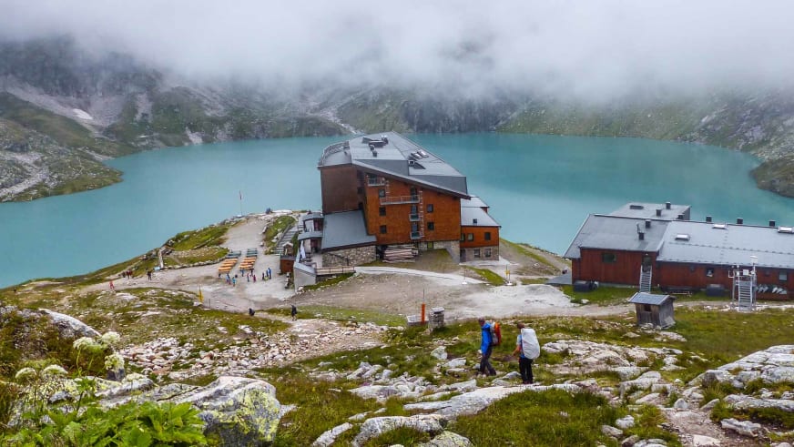 Wanderer mit Rucksäcken nähern sich dem Berghotel Rudolfshütte am türkisblauen Weißsee auf einem Bergwanderweg.
