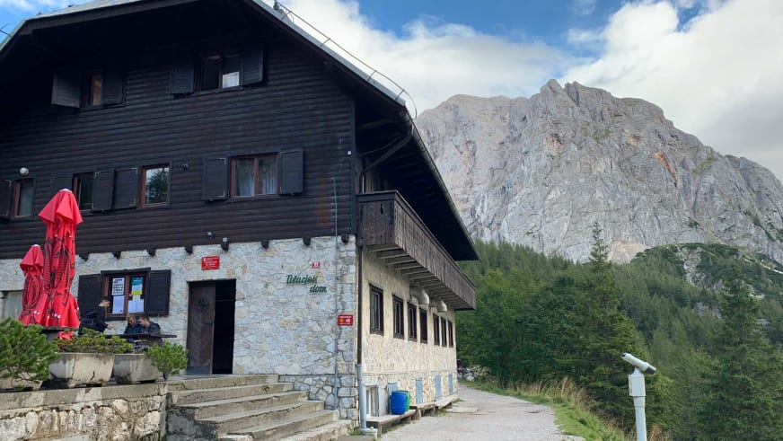 Berghaus Tičarjev dom na Vršiču vor imposanter Kalksteinwand mit Terrasse und Wegweiser.
