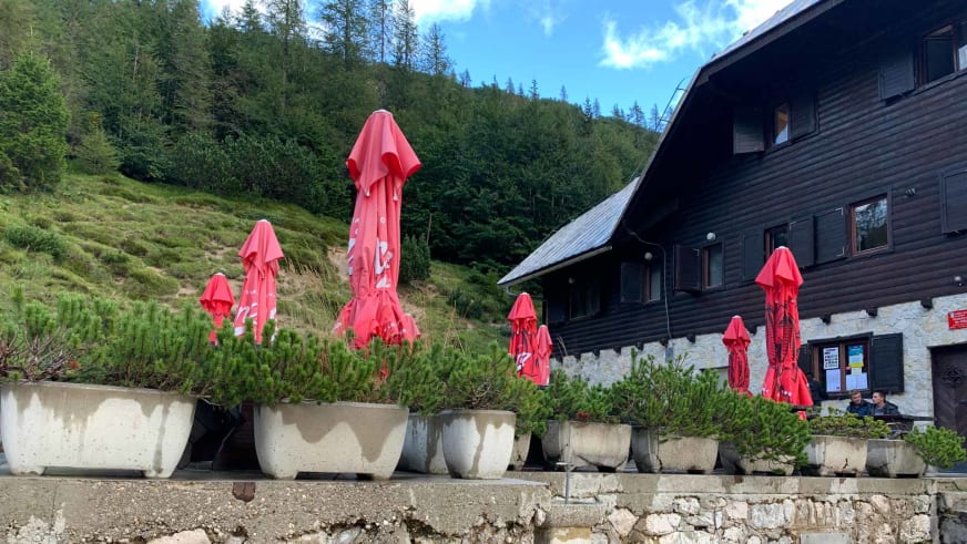 Außenbereich vom Tičarjev dom na Vršiču mit zusammengeklappten roten Sonnenschirmen vor der Berghütte.