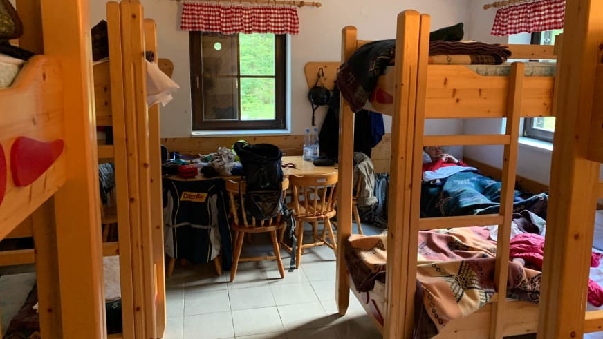 Mehrbettzimmer im Aljažev dom v Vratih mit Etagenbetten aus Holz, zentralem Tisch und rot-weiß kariertem Vorhang am Fens