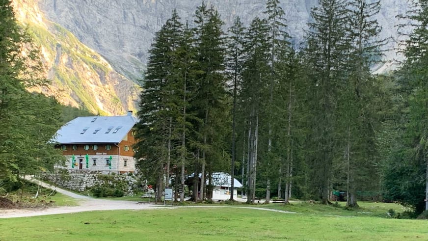 Das Aljažev dom v Vratih liegt im grünen Talkessel zwischen Nadelwald und steilen Bergflanken bei warmem Abendlicht.