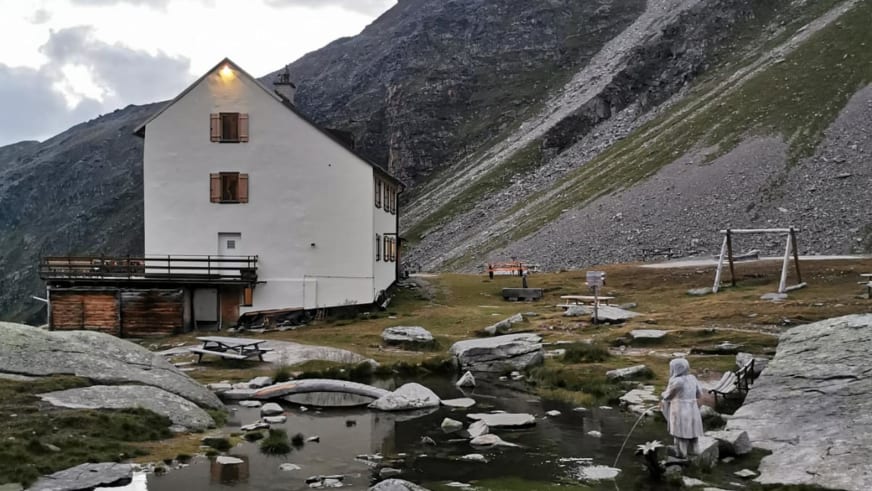Bergwanderer steht vor dem dreistöckigen Rifugio Casati mit braunen Fensterläden auf Granitplateau bei Sommerwetter.