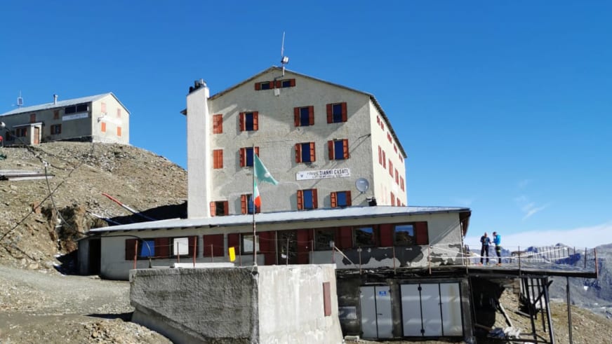 Hauptgebäude des Rifugio Casati mit hellem Stein, roten Fensterläden und italienischer Flagge vor schneebedeckten Gipfel