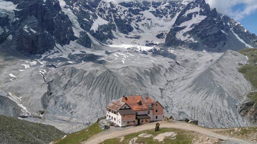 Das Rifugio Casati steht auf grüner Almwiese vor ausgedehntem Geröllfeld und schneebedeckter Gipfelkette mit Gletscher.