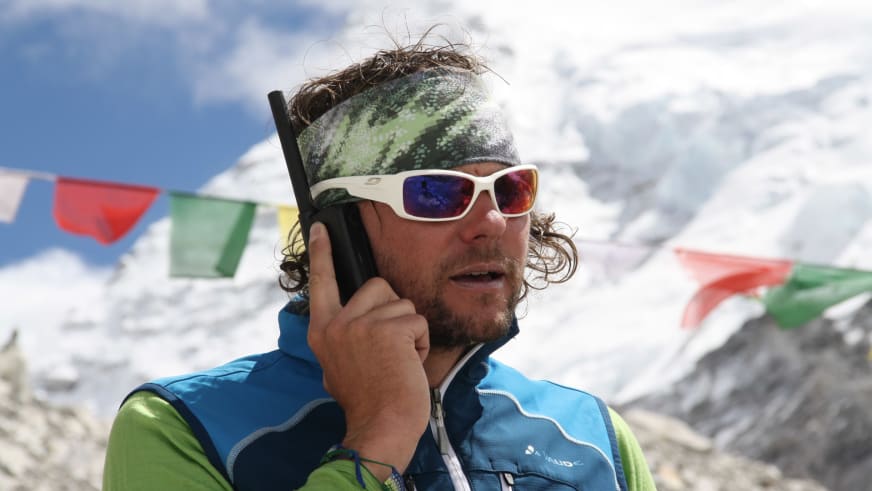 Bergführer Hans Honold mit weißer Sonnenbrille und blauer Softshell-Jacke hält ein schwarzes Funkgerät am Mund vor Gebetsflaggen.