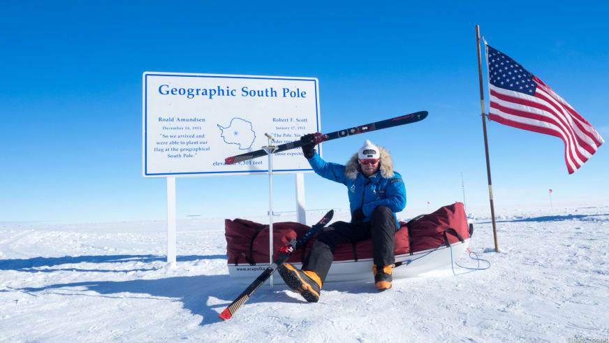Bergführer Hans Honold sitzt am geografischen Südpol auf rotem Expeditionsschlitten und hält einen Ski über dem Kopf.