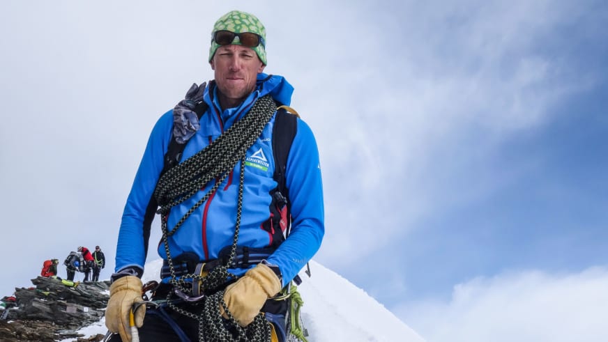 Bergführer Markus Beck steht auf schneebedecktem Berggrat mit schwarzem Klettergurt und verschiedenen Karabinern an der Ausrüstung.