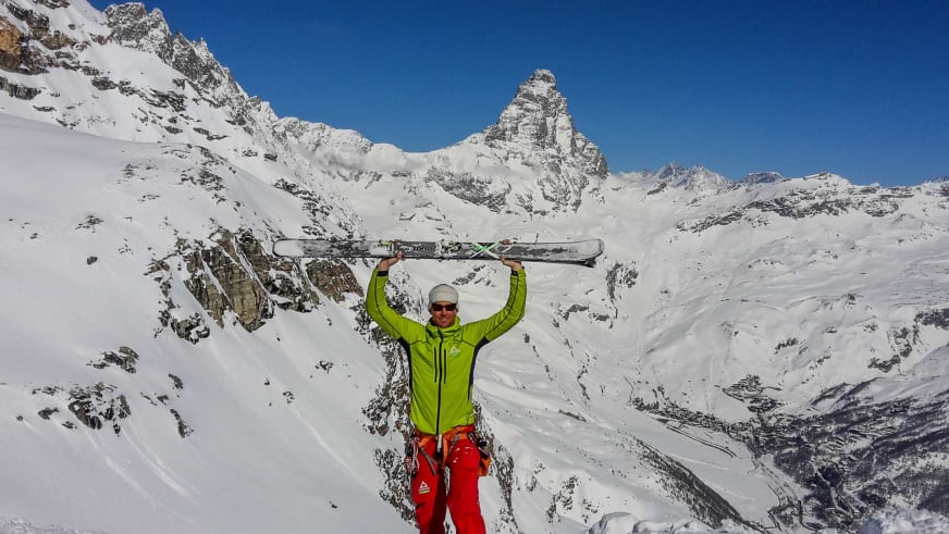 Bergführer Markus Beck hält zwei Skier über dem Kopf vor verschneiter Hochgebirgslandschaft mit pyramidenförmigem Felsgipfel.