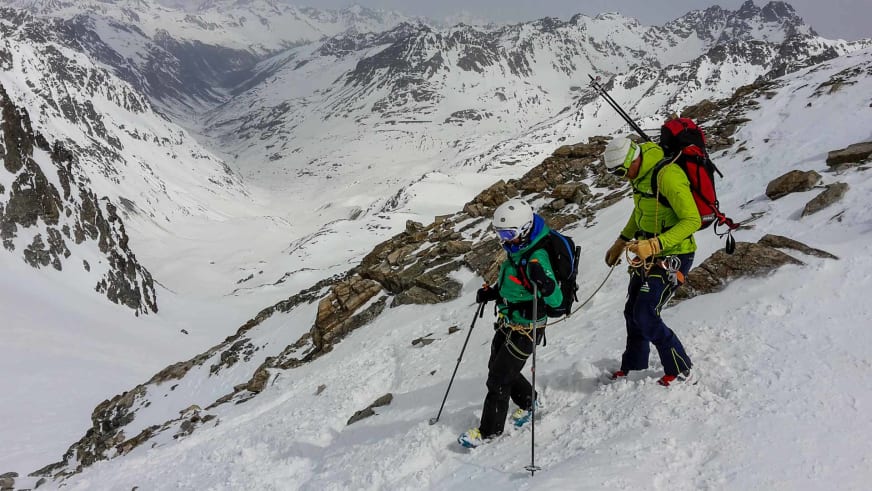 Zwei Personen mit Skitouren-Ausrüstung bewegen sich durch steiles Fels-Schnee-Gelände, angeführt von Bergführer Markus Beck in grüner Jacke.