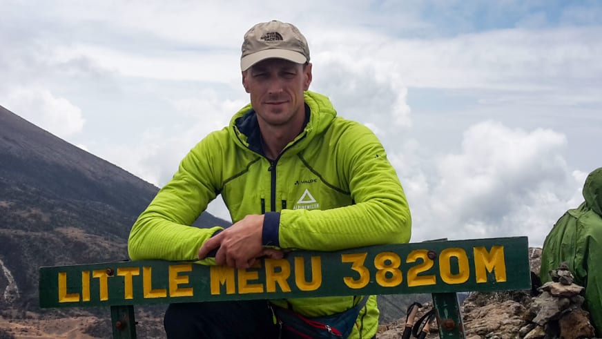 Bergführer Markus Beck steht hinter einem grünen Gipfelschild des Little Meru auf 3820 Metern Höhe in karger Berglandschaft.