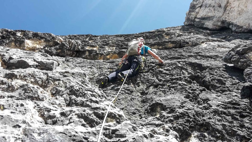 An einer steilen hellen Felswand in den Dolomiten klettert Bergführer Markus Beck mit weißem Helm und blauem Shirt.