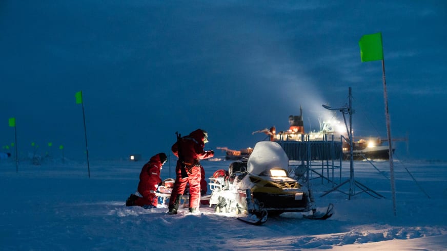 Drei Personen in roten Polaranzügen sortieren Ausrüstung bei einem Schneemobil, dabei Bergführer Markus Beck in der Polarnacht.
