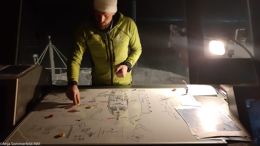 In gelbem Daunenanzug beugt sich Bergführer Markus Beck über Karten und Pläne auf einem Tisch in einem beleuchteten Innenraum.