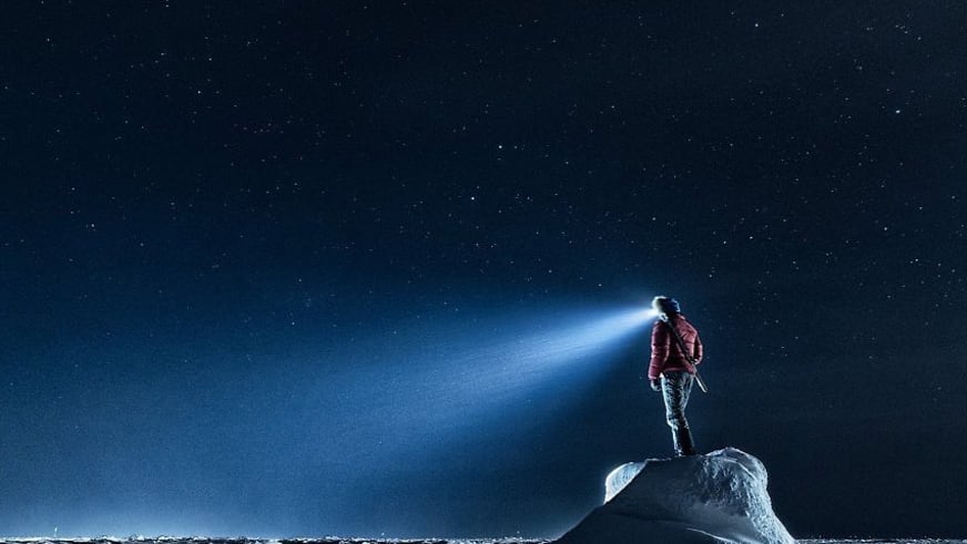 Bergführer Markus Beck steht in roter Winterkleidung auf einem Eisblock unter Sternenhimmel, die Stirnlampe leuchtet durch die Polarnacht.