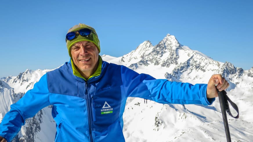 Bergwanderführer Hans Beggel lächelt vor verschneiter Bergkulisse, trägt blaue Jacke mit Alpine Welten Logo und grüne Mütze.