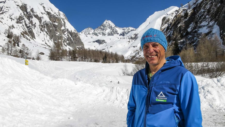 Bergwanderführer Hans Beggel mit blauer Strickmütze und Winterjacke steht lächelnd auf verschneiter Fläche vor tiefverschneitem Tal.