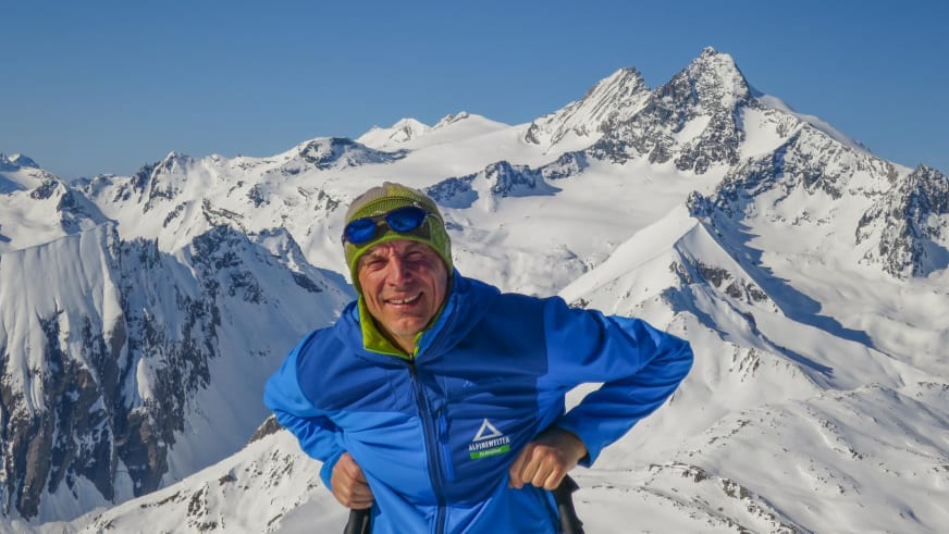 Bergwanderführer Hans Beggel hält zwei schwarze Stöcke vor verschneiter Hochgebirgslandschaft und trägt blaue Daunenjacke mit grüner Mütze.