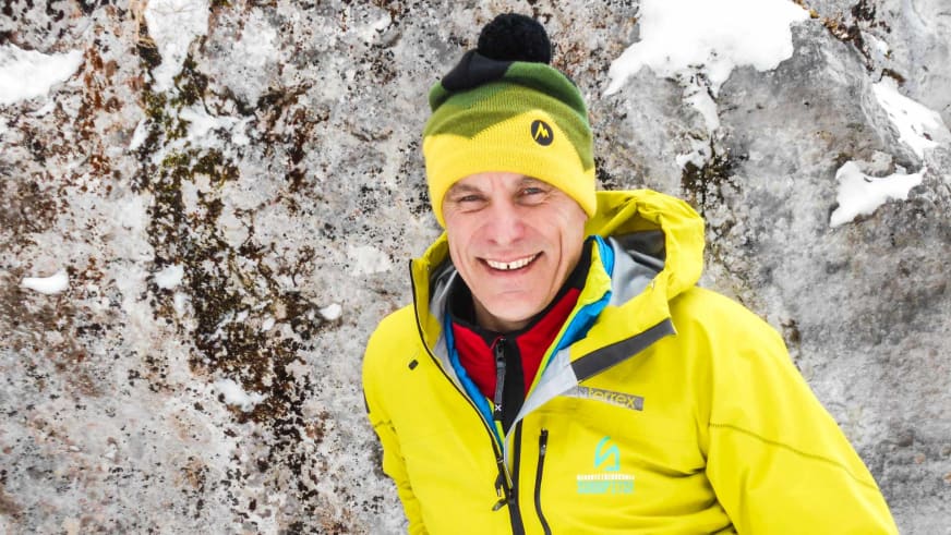 Bergwanderführer Hans Beggel lächelt vor grauer Felswand mit Schneeresten, trägt gelb-grüne Mütze und gelbe Alpine Welten Jacke.