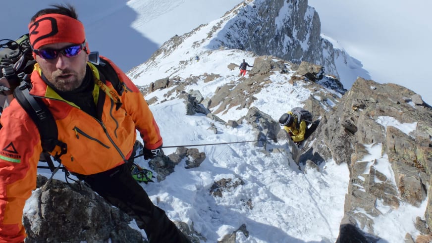 Bei einer Hochtour in der Silvretta steht Skilehrer Jürgen Brandhuber mit orangener Jacke vor verschneiter Felswand.