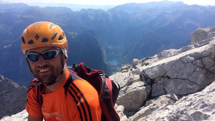 Skilehrer Jürgen Brandhuber mit orangem Kletterhelm und Sonnenbrille lächelt auf einem Gipfel vor Bergpanorama.