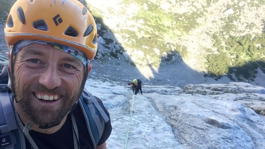 Beim Klettern macht Skilehrer Jürgen Brandhuber ein Selfie mit orangem Helm vor grauer Felswand im Sonnenlicht.