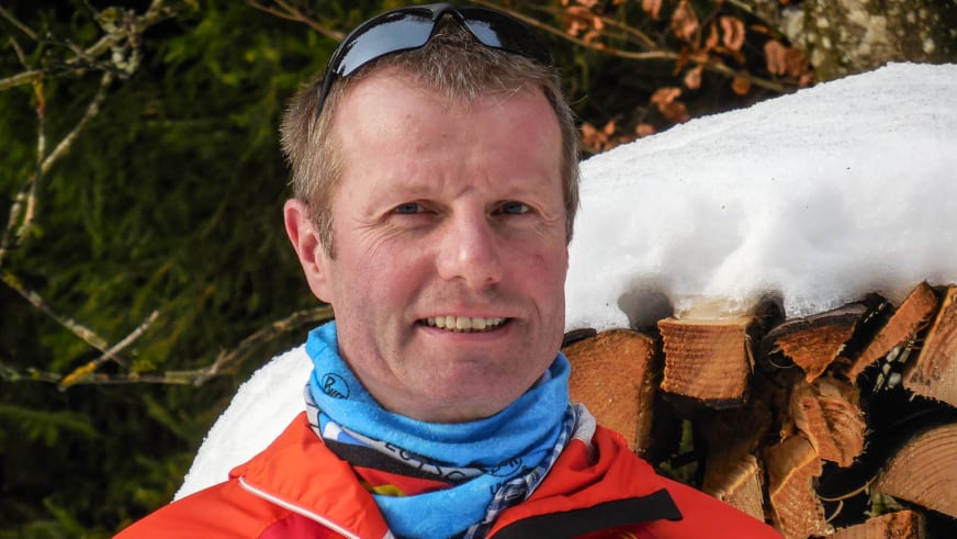 Bergwanderführer Albert Bucher in roter Softshell-Jacke und mit blauem Multifunktionstuch lächelt vor verschneitem Holzgeländer.