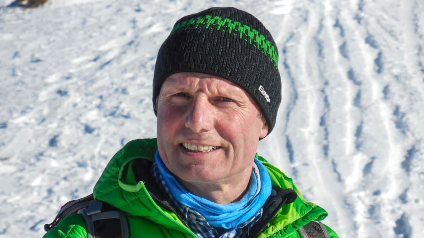 Bergwanderführer Albert Bucher lächelt in die Kamera, trägt schwarze Strickmütze und grüne Winterjacke vor Schneelandschaft.