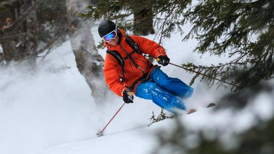 Bergführer Sebastian Conrad fährt in roter Skijacke durch tiefen Pulverschnee zwischen Nadelbäumen mit Schneepulver-Aufwirbelung.