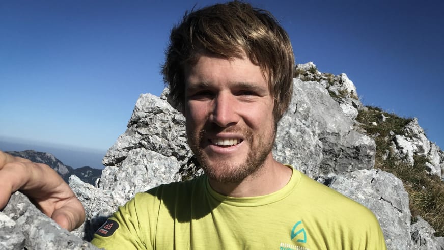 Lächelnder Bergführer Sebastian Conrad in gelbem Funktionsshirt zeigt mit der Hand nach vorne vor Kalkstein-Felswand.