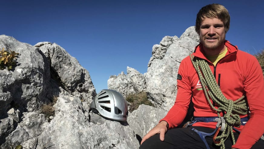 Bergführer Sebastian Conrad sitzt auf einem Felsvorsprung mit roter Softshelljacke und blauem Klettergurt.