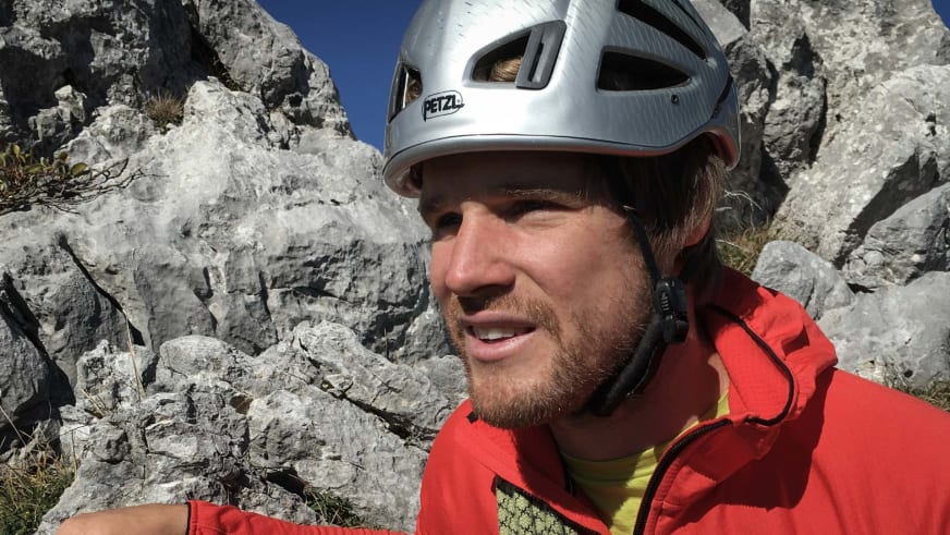 Bergführer Sebastian Conrad mit weißem Petzl-Helm und roter Bergjacke vor einer Kalkstein-Felswand.