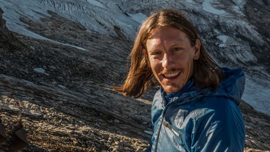 Portrait von Skilehrer Benedikt Fritz mit schulterlangen braunen Haaren und Bart in blauer Softshell-Jacke vor Felswand.