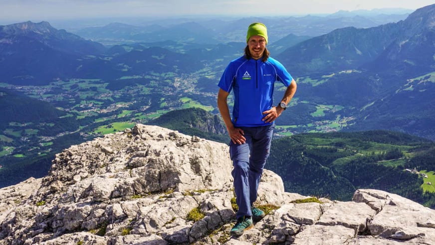 Skilehrer Benedikt Fritz steht lächelnd auf einem Felsgipfel mit grüner Mütze und blauem Funktionsshirt vor Alpenpanorama.