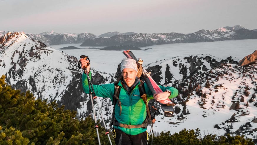 Skilehrer Benedikt Fritz trägt grüne Daunenjacke und graue Mütze, hält Tourenskier über der Schulter und blickt lächelnd in die Kamera.