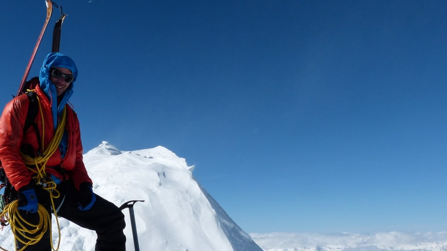 Auf einem schneebedeckten Grat steht Bergführer Bernd Budil in roter Daunenjacke mit Eispickel und gelbem Seil über der Schulter.