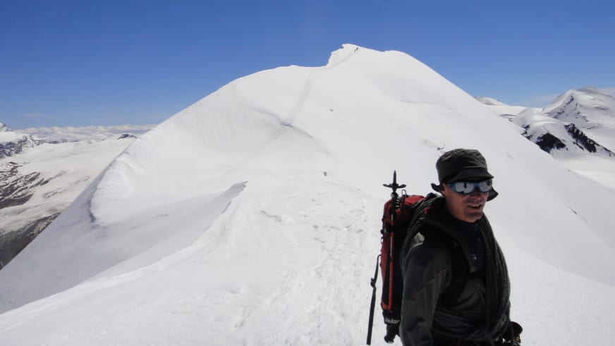 Mit Skistock und rotem Rucksack steht Bergführer Bernd Budil auf schneebedecktem Grat vor verschneiten Berggipfeln.
