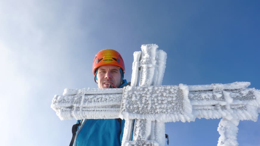 Portrait von Bergführer Bernd Budil mit orangefarbenem Helm am vollständig vereisten Gipfelkreuz bei klarem Himmel.