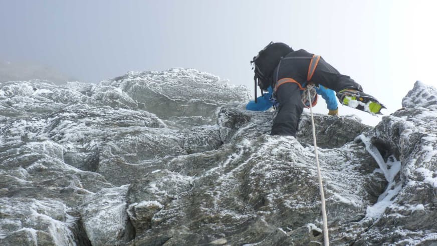 An einer vereisten Felswand klettert Bergführer Bernd Budil in dunkler Winterkleidung mit orangefarbenen Akzenten und weißem Seil.