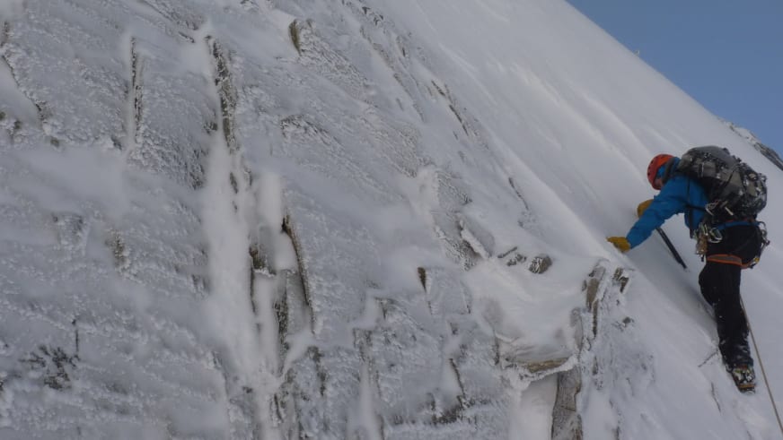 Einen steilen, schneebedeckten Felshang steigt Bergführer Bernd Budil in blauer Jacke und rotem Helm mit Bergstock auf.