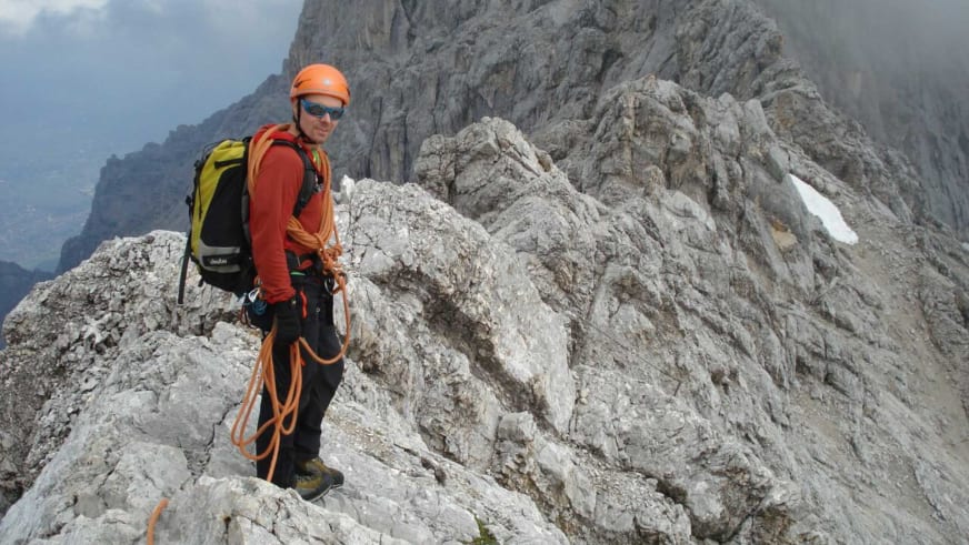 Auf einem schmalen Kalksteingrat steht Bergführer Bernd Budil mit orangefarbenem Helm, rotem Shirt und schwarzem Klettergurt.