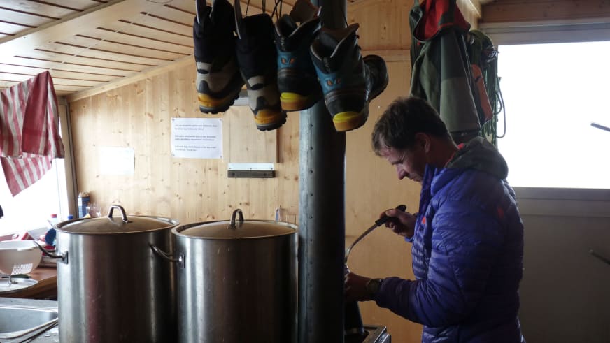 In einer Berghütte aus hellem Holz rührt Bergführer Bernd Budil in blauer Daunenjacke in einem großen Edelstahltopf.