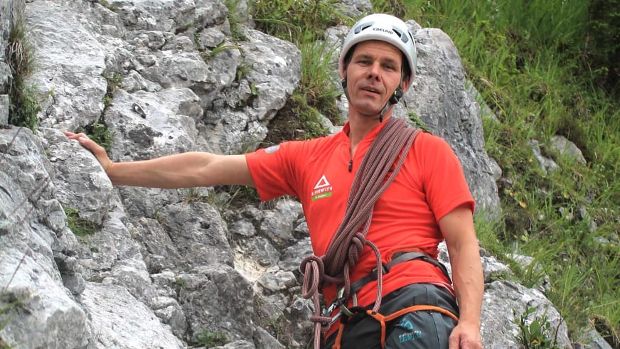 Bergführer Bernd Budil sitzt in rotem Alpine Welten T-Shirt auf einem Felsbrocken und trägt weißen Kletterhelm mit Klettergurt.