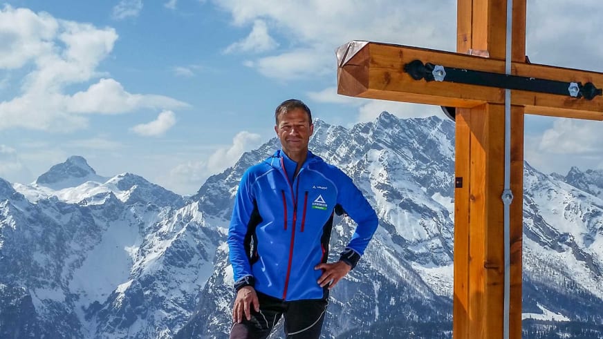 Am hölzernen Gipfelkreuz steht Bergführer Michael Höglauer in blauer Softshell-Jacke vor Panorama schneebedeckter Berggipfel.