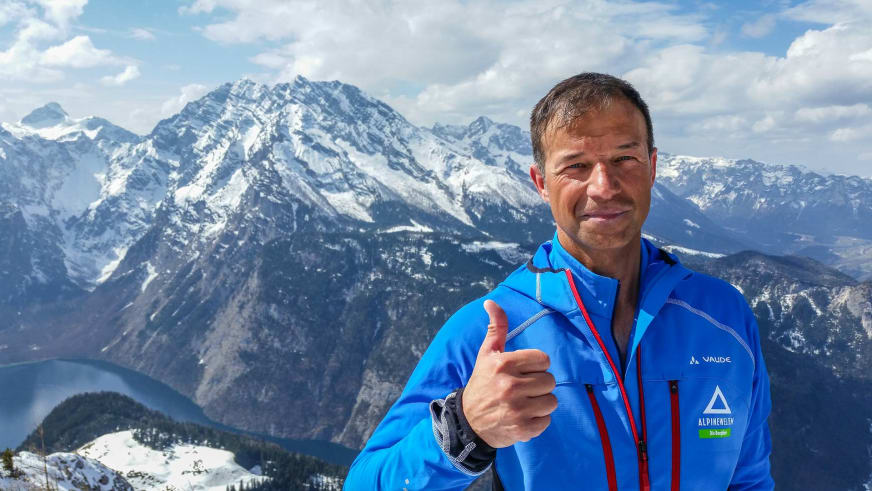 Bergführer Michael Höglauer in blauer Softshell-Jacke mit Daumen hoch vor schneebedeckten Bergen mit steilen Felswänden und bewaldeten Hängen.