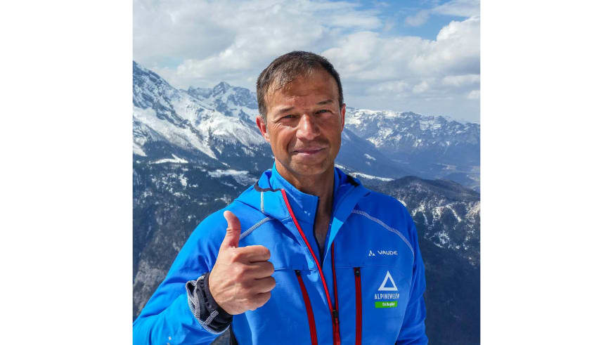 Bergführer Michael Höglauer zeigt Daumen hoch und lächelt vor schneebedeckter Gebirgslandschaft in blauer Softshelljacke.