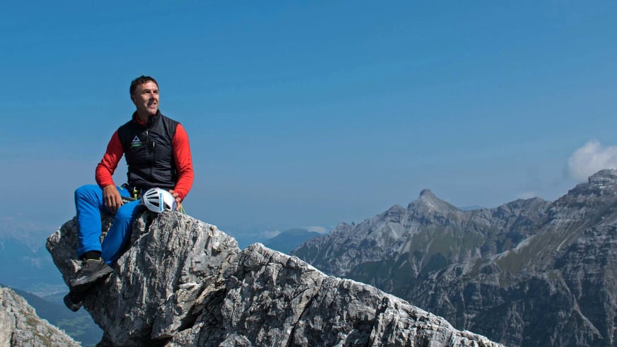 Auf einem Felsblock am Berggrat sitzt Bergführer Walter Hölzler entspannt mit weißem Kletterhelm neben sich.