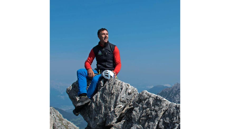 Entspannt auf einem Felsgipfel sitzt Bergführer Walter Hölzler mit weißem Kletterhelm in der Hand.