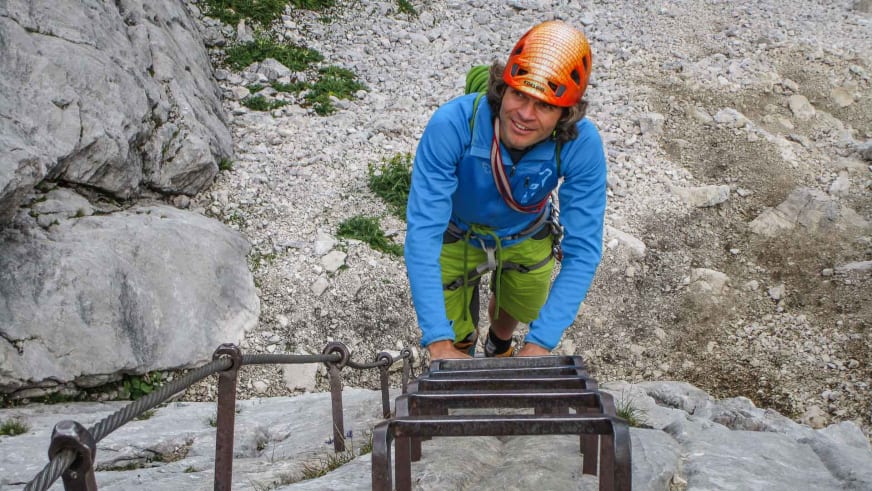 Bergführer Michael Hornsteiner in orangem Helm klettert an Stahlleiter eines Klettersteigs, gesichert am Drahtseil über Geröllfeld.