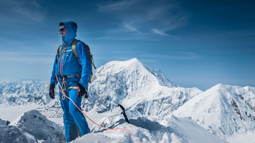 Bergführer Christoph Hummel steht in blauer Bergsteigerausrüstung auf verschneitem Gipfel vor schneebedeckter Hochgebirgslandschaft.